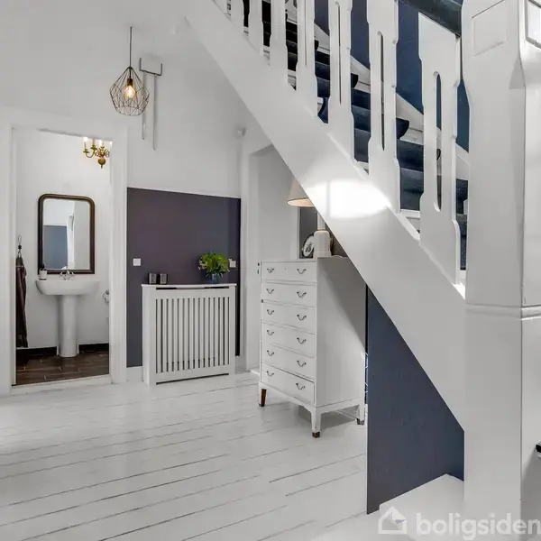 Trappe fører opad i et lyst indgangsområde med hvidmalet gulv og mørke vægge. Der er en kommode, et vægspejl og en dør til et badeværelse i baggrunden.