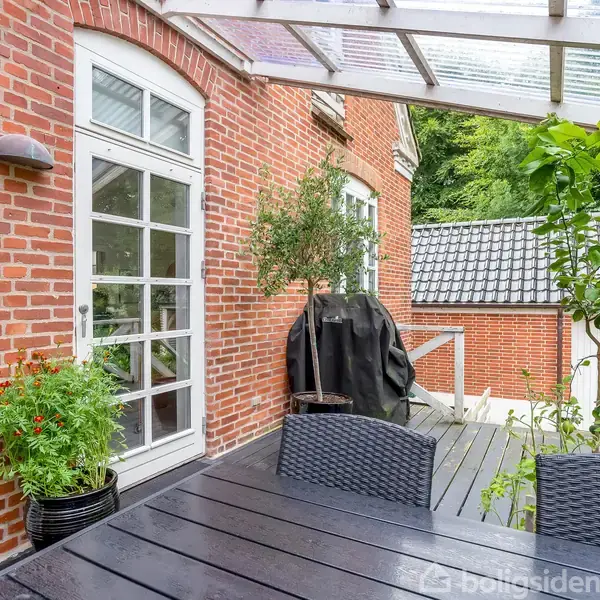 En udendørs terrasse med et træbord og flettede stole. Bordet står under en overdækket konstruktion af glas, omgivet af murstensvægge og grønne planter. En grill er dækket til.