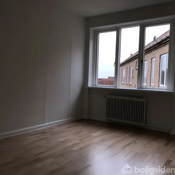 Et tomt værelse med trægulv, en radiator under et stort vindue. Udenfor ses en bygning med røde tage og flere vinduer.
