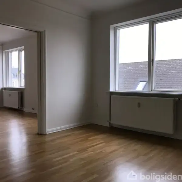 Et lyst rum med trægulv og store vinduer, der vender ud mod en murstensbygning. Radiatorer under vinduerne. Rummet synes del af en lejlighed med åbne døråbninger.