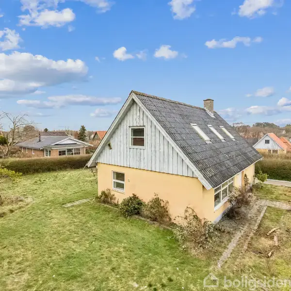 Et gult hus med en spids tagkonstruktion står i en have omkranset af hæk. Huset har flere vinduer og en skorsten. Himlen er blå med lette skyer.