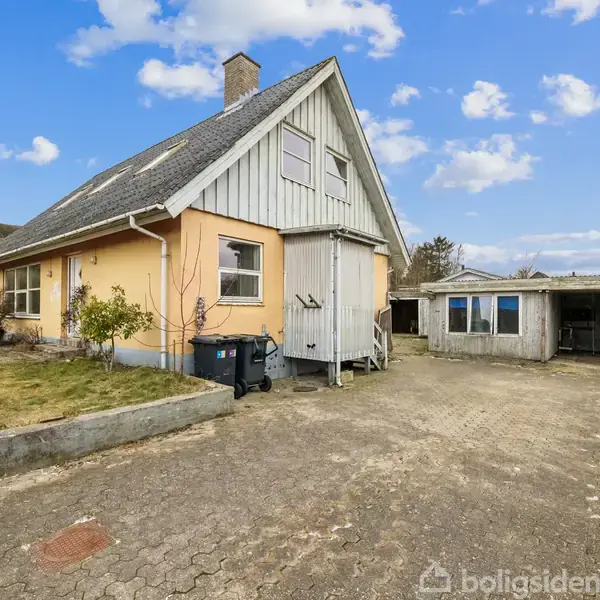 Et gult hus med gråt tag står stille ved siden af en åben garage i en flisebelagt indkørsel. Baggrunden viser en blå himmel med skyer.