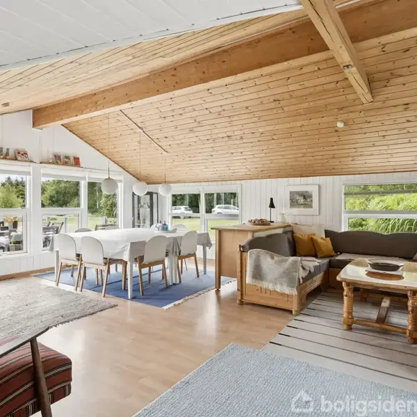 En lys stue med træloft og store vinduer. Stuen har en spisebord med stole, en sofaarrangement og forskellige tæpper på trægulvet, med udsigt til en grøn have udenfor.
