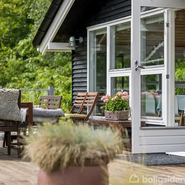 En træterrasse med stole og bord er omgivet af grøn beplantning. En potteplante med lyserøde blomster står ved døren til et sort træhus.