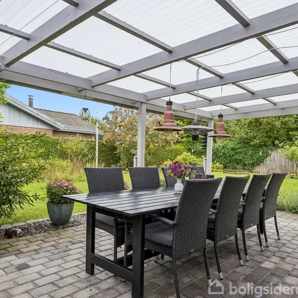 Et langt, sort havebord med otte fletstole står under en overdækket terrasse. To røde pendellamper hænger over bordet. Haven er grøn med buske og et plankeværk i baggrunden.