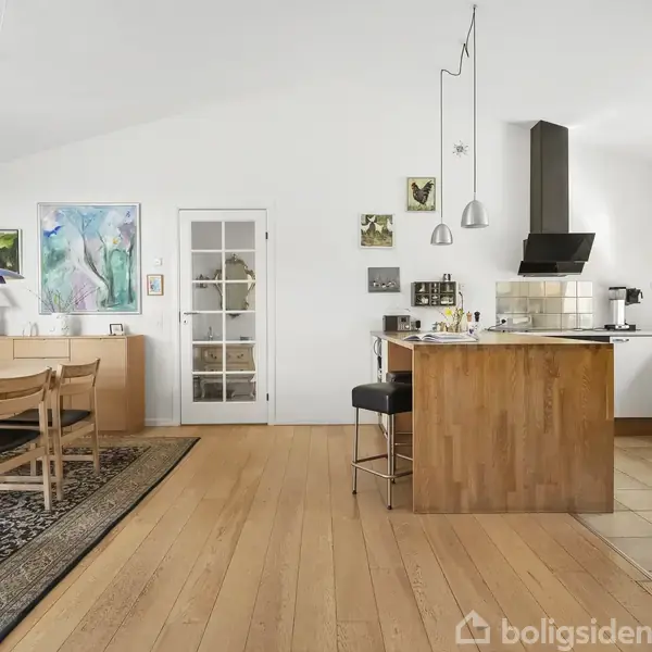 Et moderne køkken-alrum med træbord og stole, lyse lamper hængende over bordet, og køkkenø med barstole. Lyst trægulv og kunst på væggene i et rummeligt, lyst rum.
