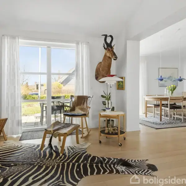 Værelse med en stor vindue indrettet med en mønstret tæppe på trægulvet, en antilopebuste på væggen, stole, et bord med planter, og en tilstødende spisestue bagved.