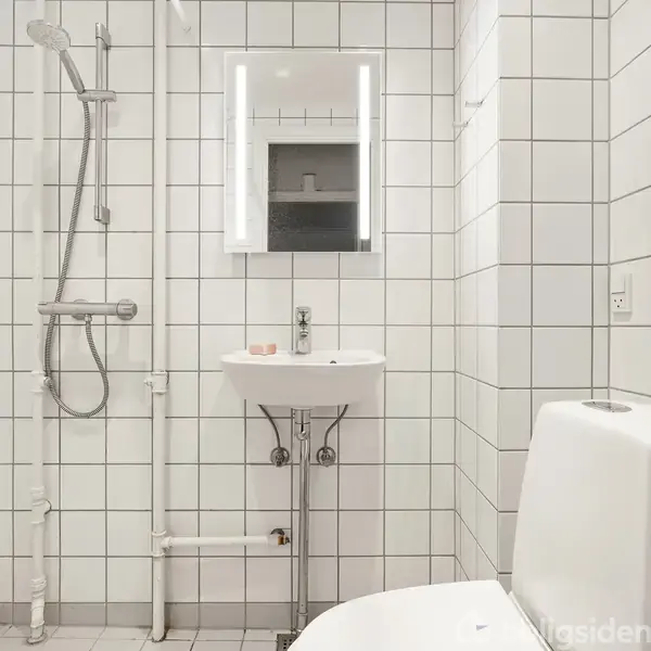 Hvidt flisebeklædt badeværelse med bruser til venstre, håndvask under spejl i midten, og toilet til højre. Enkel og funktionel indretning uden tekst.