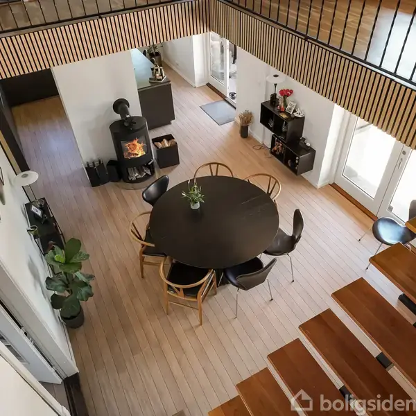 Fra oven ses en spiseplads med et rundt bord og seks stole. I baggrunden brænder en brændeovn ved siden af glasdøre, der lukker lys ind i rummet.