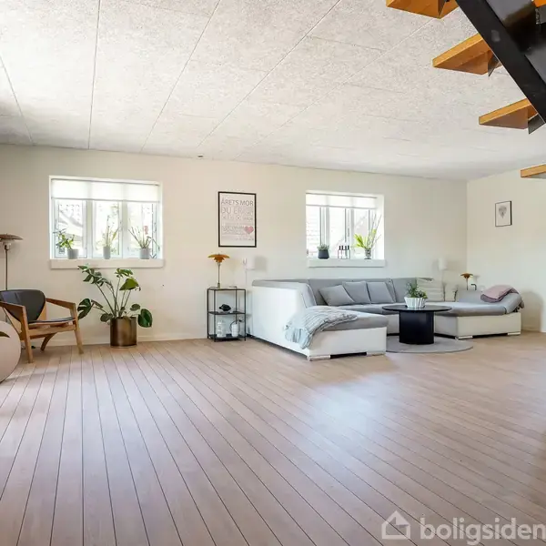 En moderne stue med en grå sofa og et rundt sofabord. En trappe fører til en anden etage. Rummet er lyst med planter og kunst på væggene.