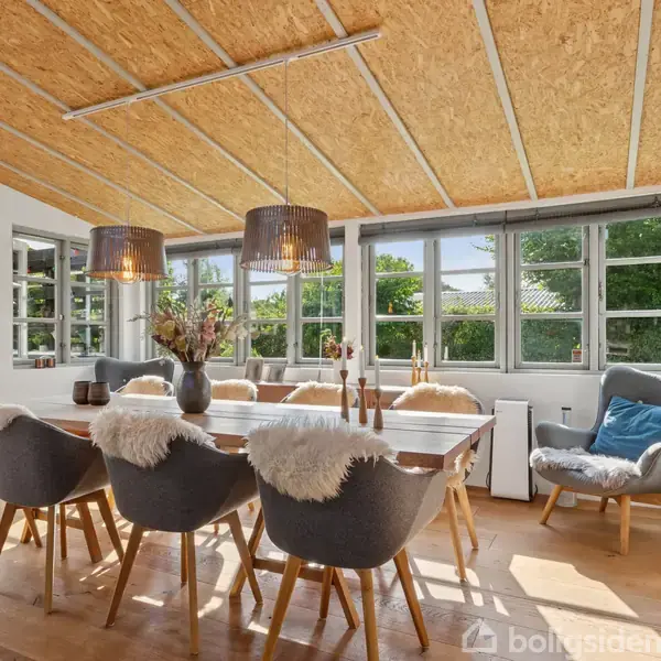 Et stort bord med grå stole og fåreskind ligger i et lyst rum med trægulv og træloft. To lamper hænger over bordet, omgivet af store vinduer med udsigt til en have.