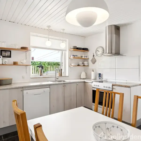 Et lyst køkken med et spisebord med træstole i forgrunden. Køleskabet står til venstre, og over bordpladen hænger hylder med køkkenudstyr. Vindue med udsigt til grøn have.