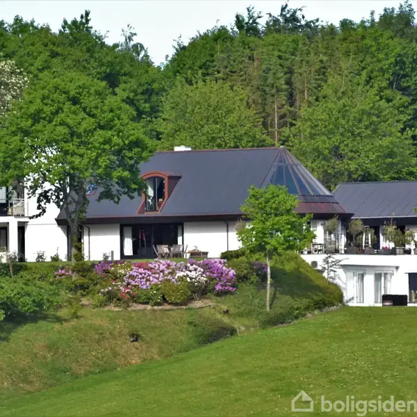 En stor hvid villa ligger på en grøn bakke omgivet af træer og buske med blomster. Huset har flere etager og et skråt tag med et glaspyramideformet udbygning.