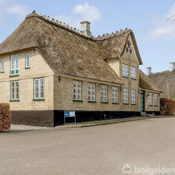 Stråtækt hus med vinduer og skorstene står langs en vej. Skilt ved siden af huset læser: "Cykelsti forbudt." Den omkringliggende hæk er veltrimmet.