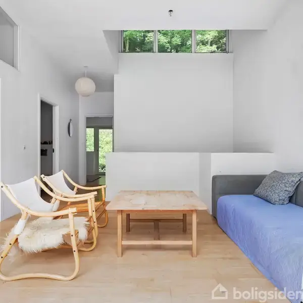 Sofa dækket med blå tæppe står ved siden af et træbord i lys stue med to stole. Store vinduer ved loftet giver udsigt til grøn bevoksning.