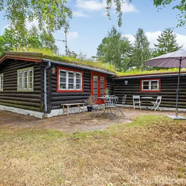 Træhus med græsbeklædt tag står stille i en grøn have. Flere havestole og et bord med parasol pryder fliseterrassen. Omgivelserne er fyldt med træer.