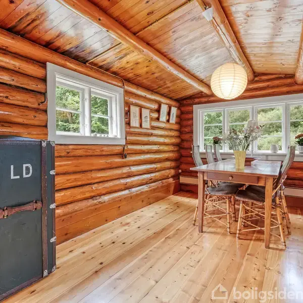 En træbeklædt spisestue indeholder et træbord med fire stole. En sort kuffert mærket "LD" står op ad væggen. Flere vinduer giver udsigt til naturen udenfor.