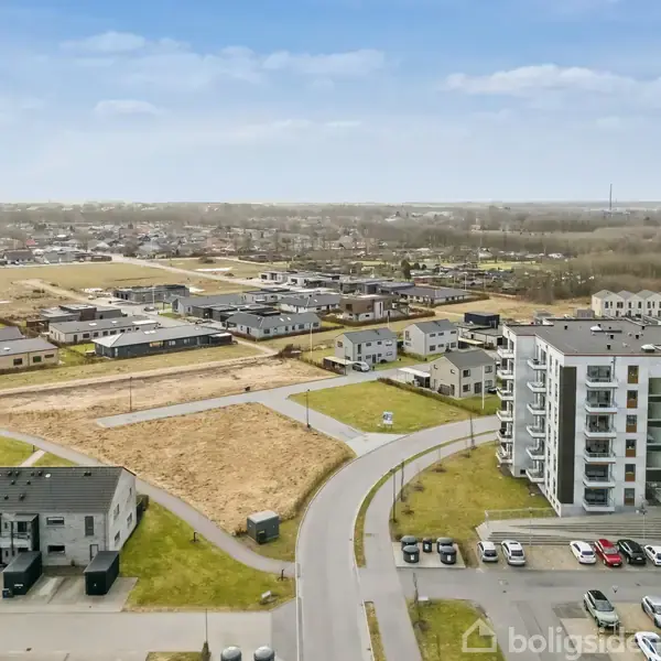 Et moderne lejlighedskompleks står ved siden af en parkeringsplads, omgivet af åbne marker og flere lavere bygninger i et forstadsområde. Klare blå himmel på en solrig dag.