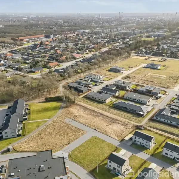 Boligområde set fra luften. Nyopførte huse ligger på parallelle rækker med grønne plæner og ubebyggede grunde ved siden af. I baggrunden ses en by med flere bygninger.