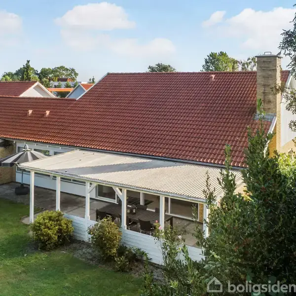 Huset med rødt tag og skorsten står stille i en frodig have. Der er en overdækket terrasse foran huset, omgivet af grønne buske og græsplæne.