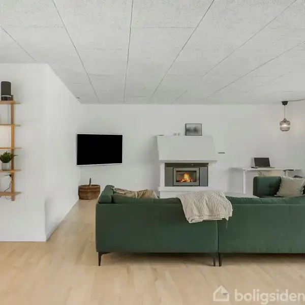 En grøn sofa står i en moderne stue med lyst trægulv. Et vægmonteret TV hænger ved siden af en pejs. Et lille arbejdsområde ses i baggrunden ved et stort vindue. En åben hylde med planter og bøger er placeret til venstre.