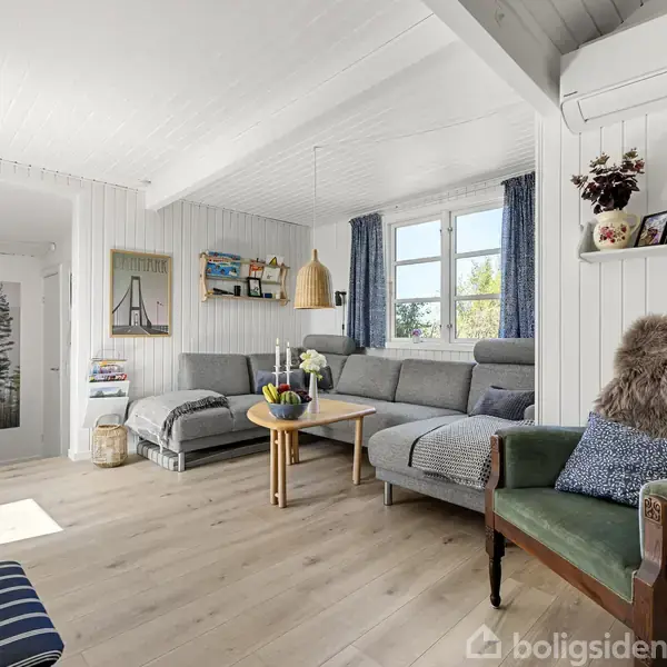 Stue med grå hjørnesofa og træbord optager rummet. Grøn lænestol med pude og tæppe står til højre. Store vinduer slipper lys ind. Billede af skov ses i baggrunden.