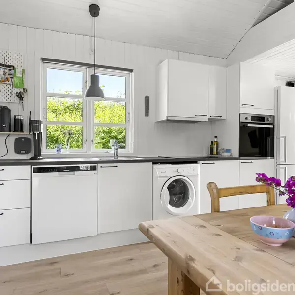 Et lyst køkken indeholder hvide skabe, vaskemaskine og indbygningsovn. En træbord står med lilla blomster i en vase. Et vindue giver udsigt til en grøn have.