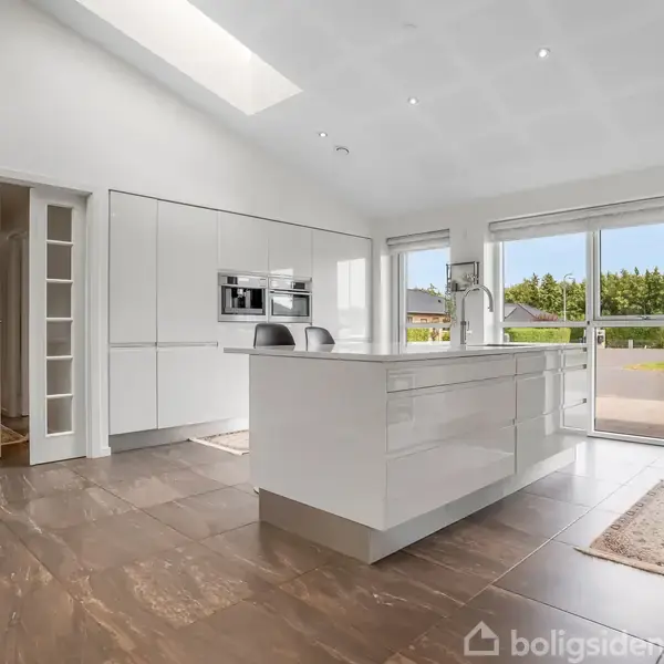 Et moderne køkken med en stor, hvid ø i midten. Køkkenet har hvide skabe, åbne glasdøre og store vinduer, der giver udsigt til en terrasse med grønne områder udenfor.