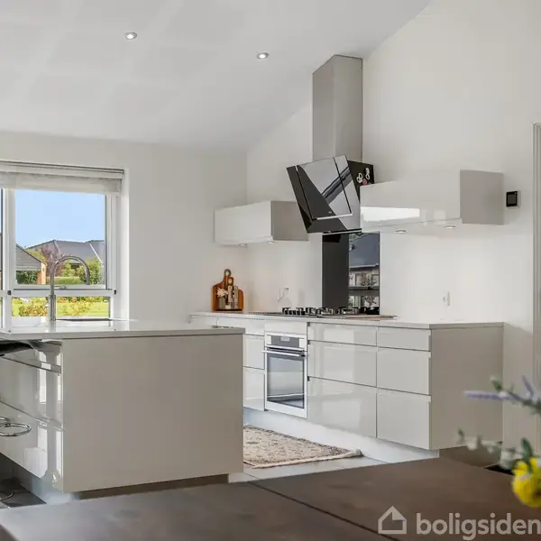 Et moderne køkken med en ø i midten, to høje stole, gaskomfur og emhætte. Store vinduer giver udsigt til en have udenfor. Blomster står i forgrunden.