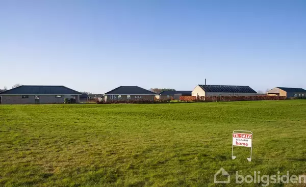 Græsplæne med "Til Salg" skilt, omkranset af moderne huse, i en forstadsområde. Klar blå himmel danner en rolig baggrund. Skilttekst: "Til Salg 52 40 31 08 home".