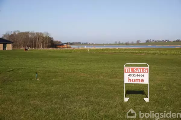 Et skilt i græs med teksten "TIL SALG 65 32 44 04 home" i forgrunden. I baggrunden ses en sø og skovområde.