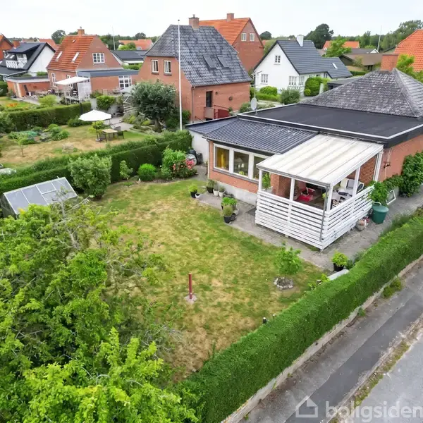 Hus med sort tag og veranda omgivet af en velplejet have med hække og træer. Drivhus ses til venstre, beliggende i et boligområde med lignende huse.