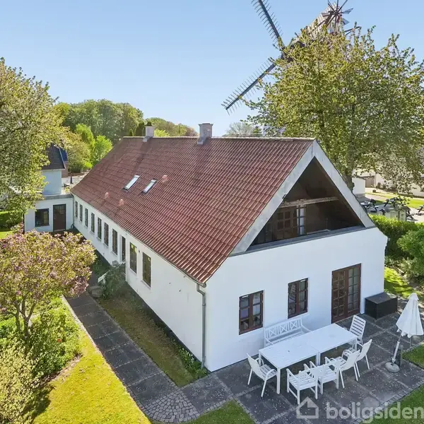 En hvid, traditionel bygning med rødt tag står i en frodig have. I forgrunden er et hvidt havemøbelarrangement. I baggrunden ses en vindmølle og flere træer.