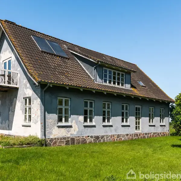 Et hvidt, to-etagers hus står med en altan og solpaneler på taget. Det er omgivet af en velplejet græsplæne og træer under en klar blå himmel.