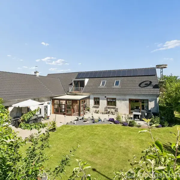 Et hus med solpaneler på taget står midt i en grøn have. Terrassen er udstyret med parasol og møbler, omgivet af frodig vegetation og en klar blå himmel.