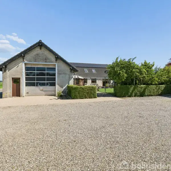 Bygning med en stor garageport står på en grusbelagt gårdsplads, omgivet af grønne buske og træer. Bagved er der et stort hus med sorte tagsten og flere vinduer.