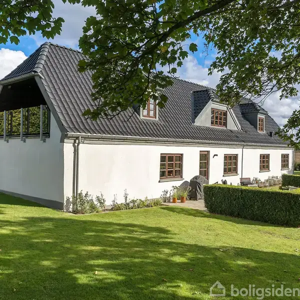 Hvidmalet hus med mørkt tegltag står i en velplejet have med hække og en lille terrasse. Store træer omgiver ejendommen, og himlen er skyet.