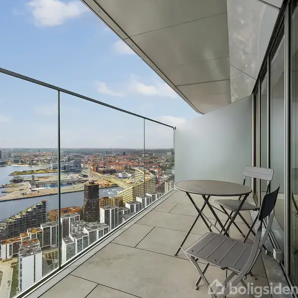 En moderne altan med glasrækværk har et rundt bord og to stole. Udsigt over en havn med moderne bygninger og bylandskab i baggrunden, under en klar himmel.