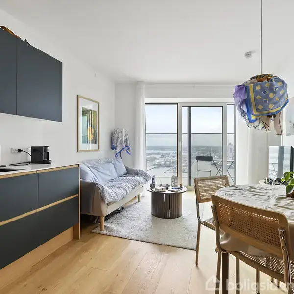 Et moderne køkken-alrum med mørke skabe og integreret vask står ved siden af en spiseplads. Et sofahjørne med grå sofa leder ud til en balkon med udsigt.