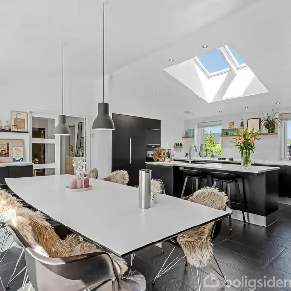 Et moderne køkken med lang spisebord fyldt med pelse, og hængende lamper. I baggrunden er et køkkenområde med sort kabinetter, ø og ovenlysvindue, omgivet af planter og dekorative elementer.
