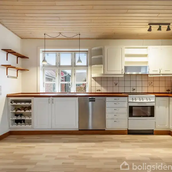 Køkken med hvide skabe og træbordplade, opvaskemaskine, komfur og åbne hylder. Rummet har lyst trægulv og et vindue, der lukker naturligt lys ind, hvilket skaber en indbydende atmosfære.