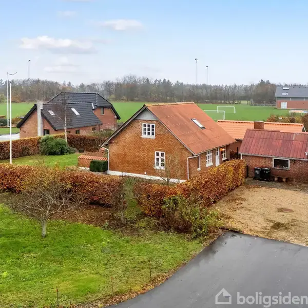 Et rødt murstenshus ligger mellem hække i et grønt boligområde. Nabohuse står tæt langs vejen, og en sportshal ses i baggrunden. Grusbelagt garageområde er foran huset.