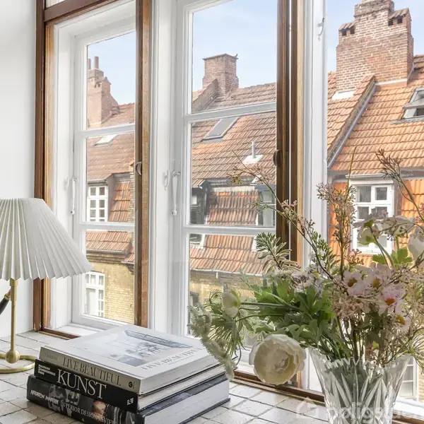 Vask i et lyst køkken med træbordplader, under et åbent vindue med to lamper. Udenfor ses et tegltag og blomster. Hylder og skabe er på væggen til højre.