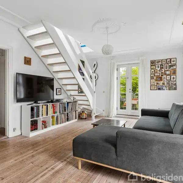 Stue med grå sofa, tv på væggen, trappe til venstre, åbent mod en dør til et badeværelse, hylder med bøger og en billedvæg. Lyse trægulve og franske døre.