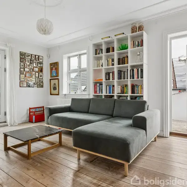 En grå sofa står i en lys stue med trægulv. Ved siden af er en bogreol fyldt med bøger. Store vinduer og en åben dør leder til en altan. Væggen har mange billedrammer.