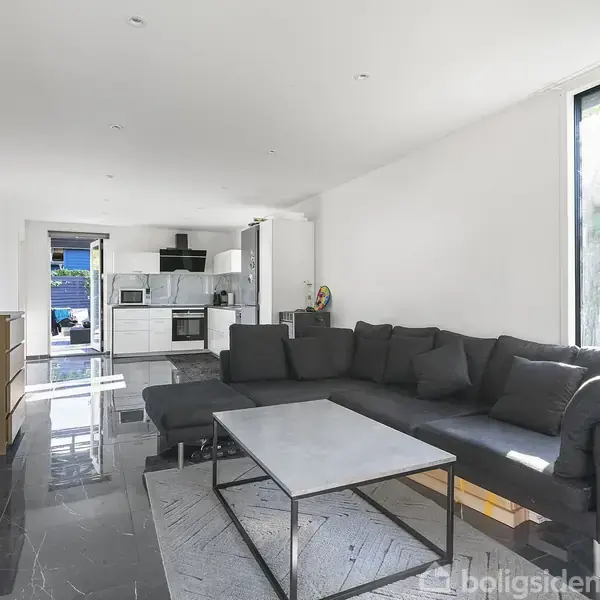 Sort sofa står i en moderne stue med gråt marmorgulv; den vender mod et kvadratisk sofabord. Et åbent køkken med hvide elementer ses bagved, badet i naturligt lys.