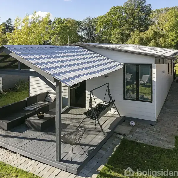 Et moderne, hvidt sommerhus med overdækket terrasse, hvor en hængesofa og sorte udemøbler står. Omgivet af grøn beplantning på en solrig dag.
