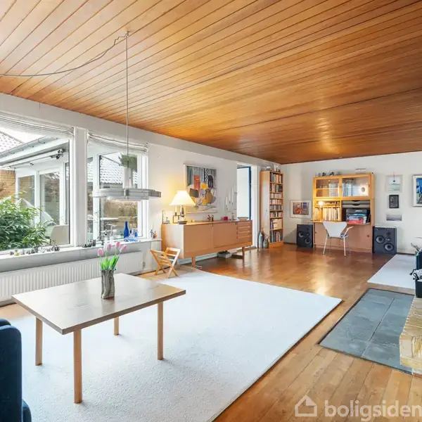 Stue med et træbord på et lyst tæppe, omgivet af blå sofa og pejs. Store vinduer giver udsigt til haven, og lyse træmøbler står langs væggene. Ingen tekst fundet.