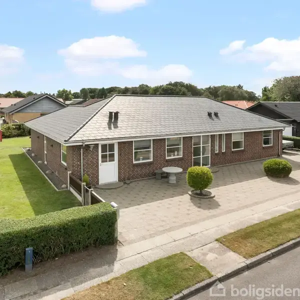 En murstenshus står stille i en velplejet have med grønne buske og en flagstang med Dannebrog. Huset har grått tag og store vinduer.