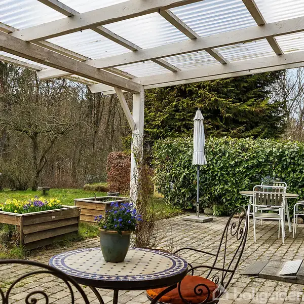 Terrasse med møbler under gennemsigtigt tag. Rundt bord med blomsterkrukke foran metalstol. I baggrunden ses græsplæne, planter, grøn hæk og skov. Markerede fliser dækker gulvet.